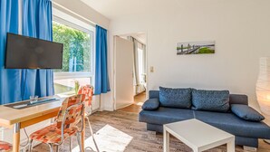 Uma sala de estar moderna com televisão de tela plana, sofá, mesa de jantar com cadeiras e vista para o exterior através de grandes janelas.