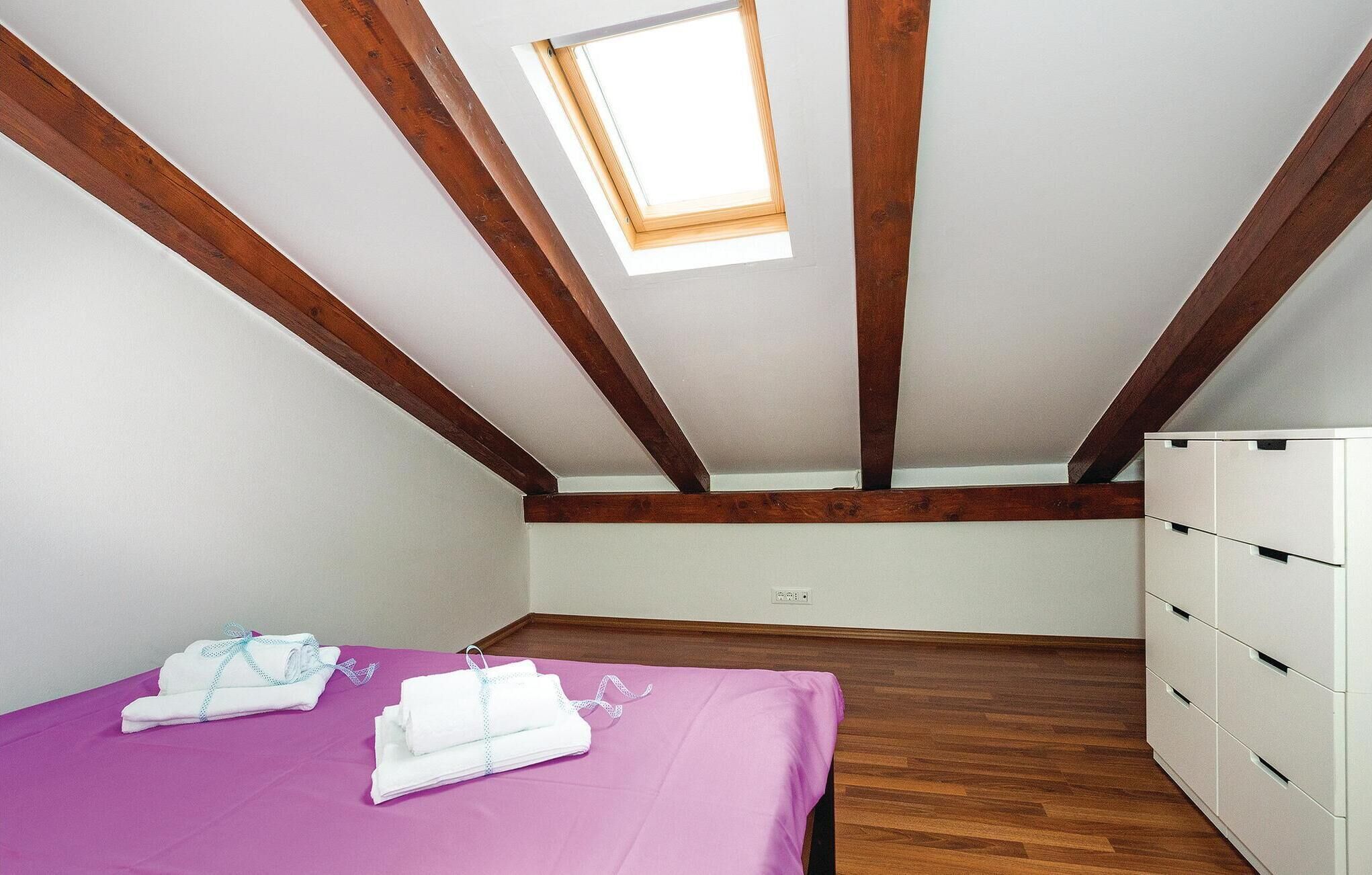Een kamer met een schuine balkenplafond, een dakraam, een bed met paars beddegoed en een witte kapstok.