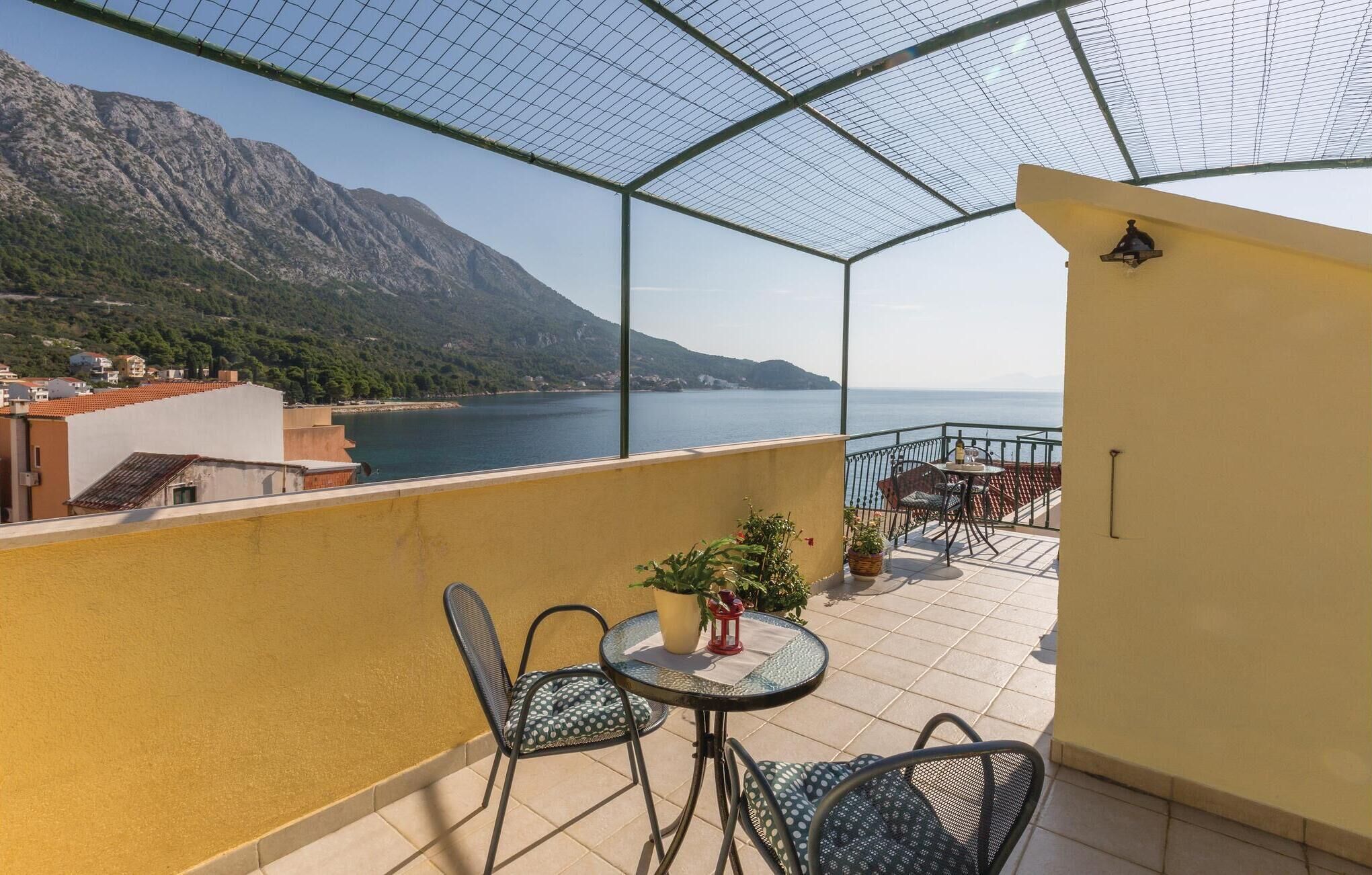 A balcony with a view of a coastal landscape, including mountains and a body of water. There are two chairs and a small table with a potted plant.