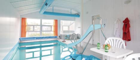 An indoor swimming pool with a blue inflatable shark, a white table with drinks, and a window with orange curtains.