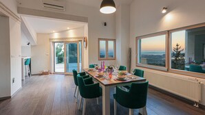 A dining area with a table set for four, green chairs, a view of the city through large windows, and a sliding glass door leading to a balcony.