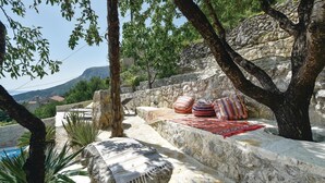 A stone bench with cushions and a blanket, surrounded by trees and a stone wall.