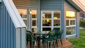 A covered patio with a table and chairs, large windows, and a view of the sunset. - Friedrichskoog-Strandpark 15 (Friedrichskoog)