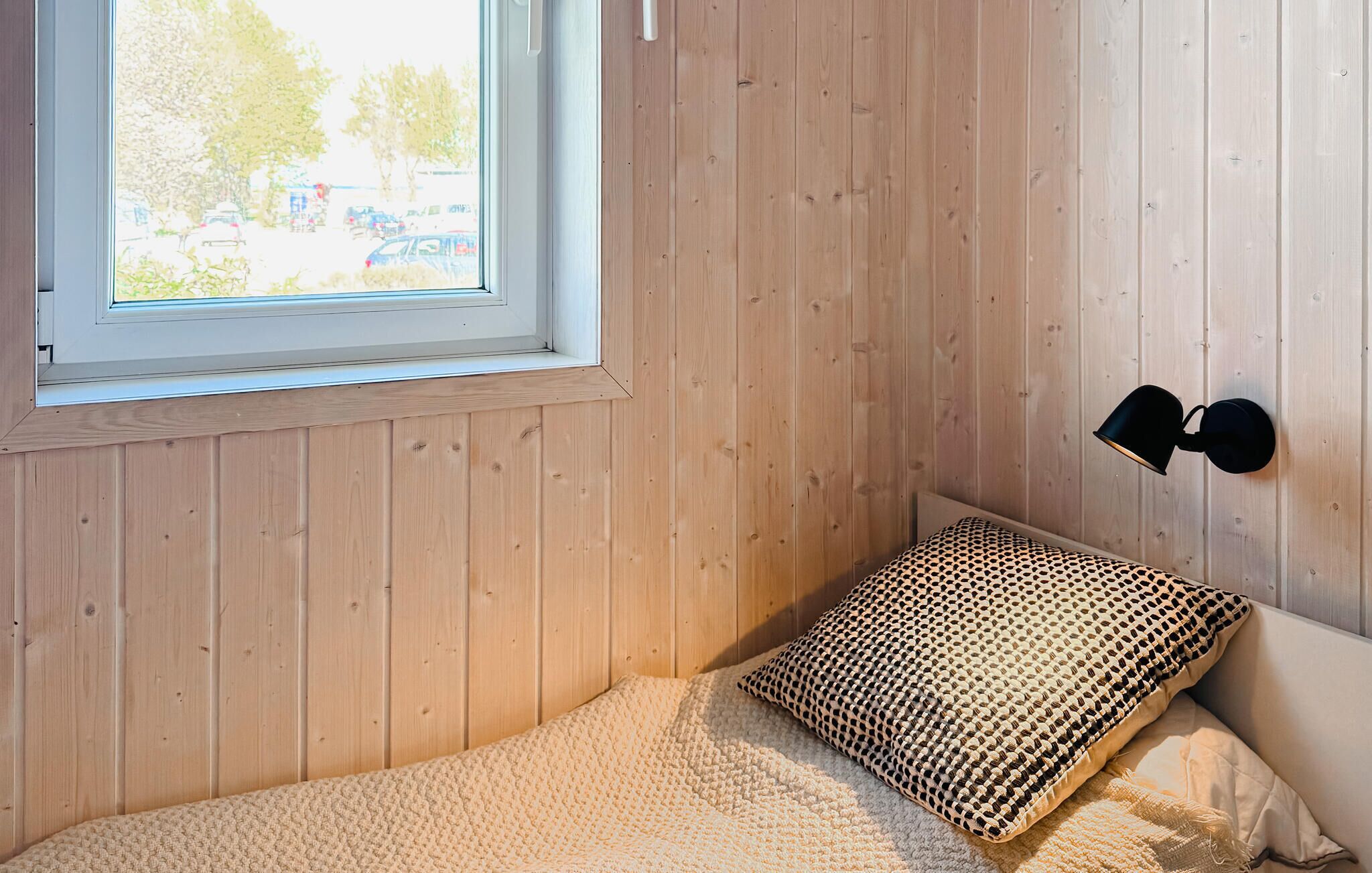Un dormitorio con ventana, paredes de madera, una cama con almohada estampada y una lámpara fijada a la pared.