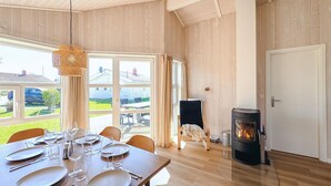 Un coin repas avec une table dressée pour quatre personnes, un poêle à bois noir et une vue sur l’extérieur grâce à de grandes fenêtres.