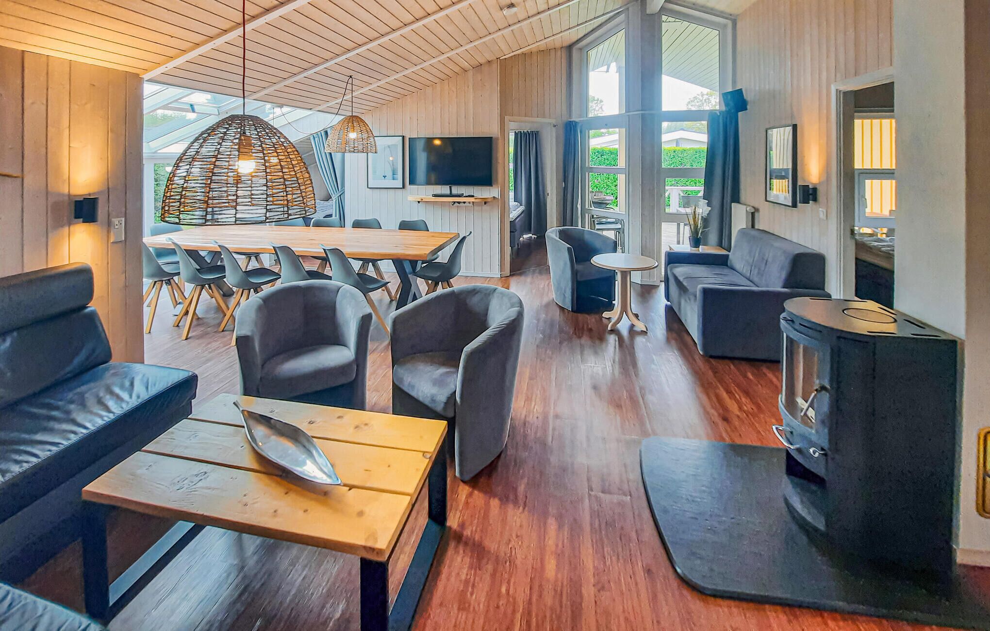 A modern interior with a wooden ceiling, a large dining table, grey sofas, and a wood-burning stove.