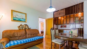 A cozy living room with a brown leather sofa, a blue checkered throw, a wooden dining table with chairs, a painting on the wall, and a kitchen in the background.