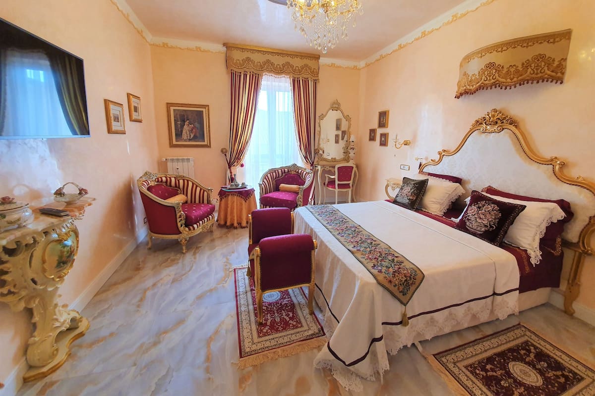 Um quarto luxuoso com uma cama grande, duas poltronas, uma toucador com espelho e um candelabro.