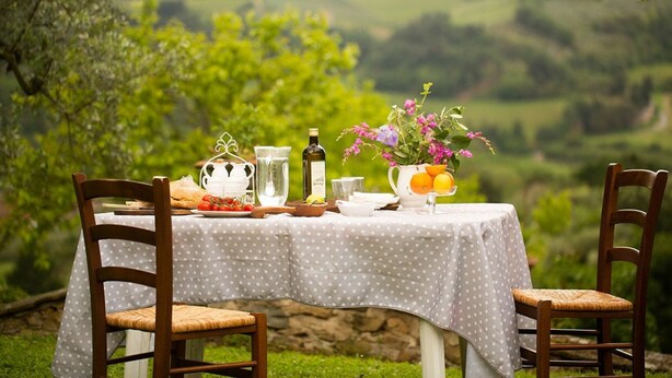 Una mesa al aire libre con mantel estampado de lunares, rodeada de sillas y varios objetos como una tetera, una botella de agua y fruta.