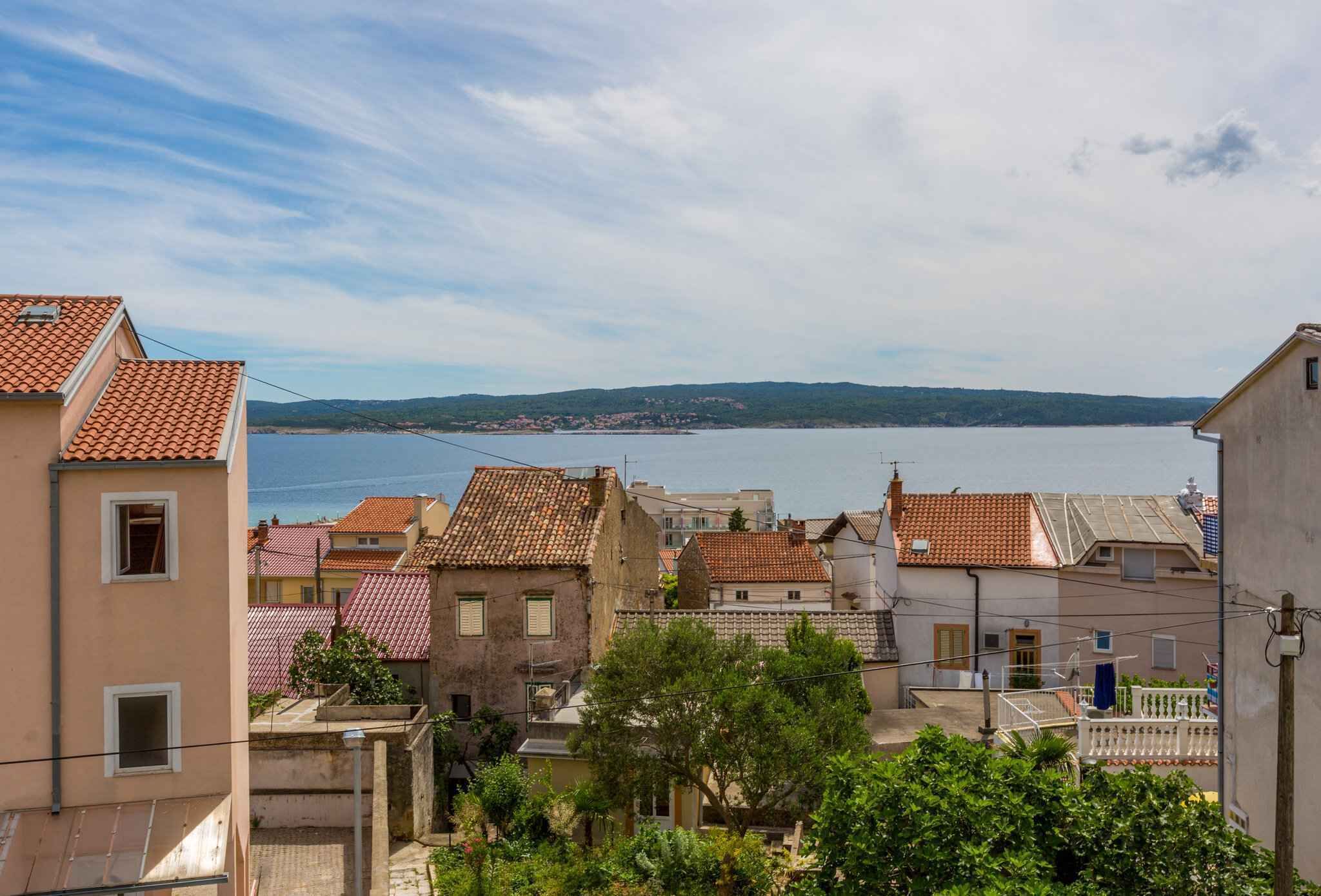 A coastal town with terracotta-roofed buildings, a view of the sea, and a hilly landscape in the distance.