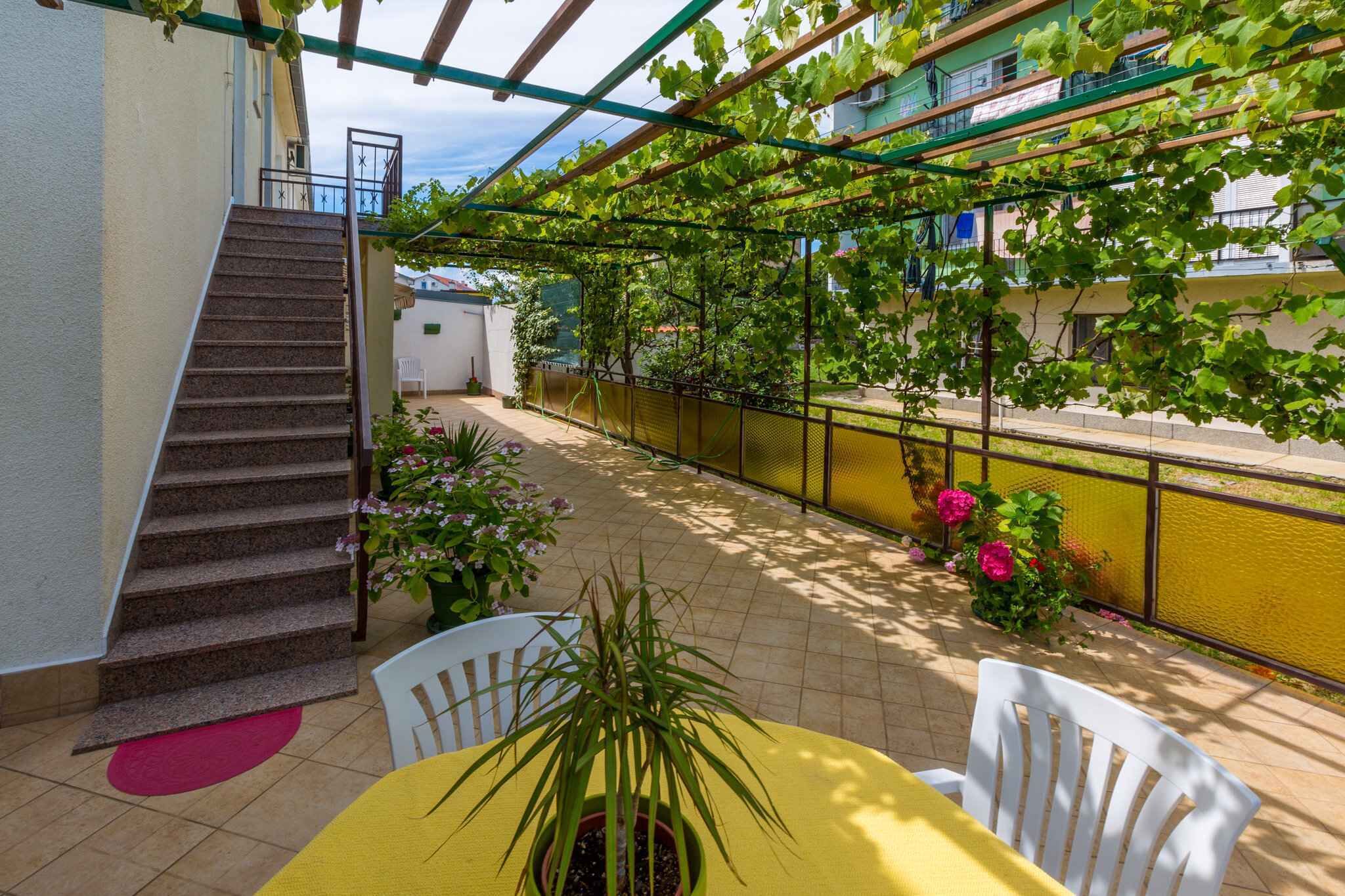 A covered outdoor area with a staircase, potted plants, and a yellow table with white chairs.