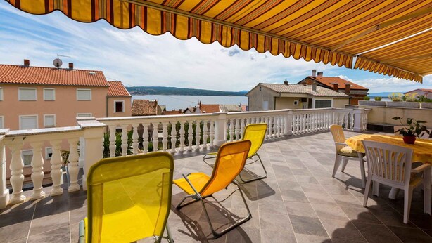 Eine Terrasse mit gelben Stühlen und einem Tisch, mit Blick auf ein Gewässer und Gebäude.