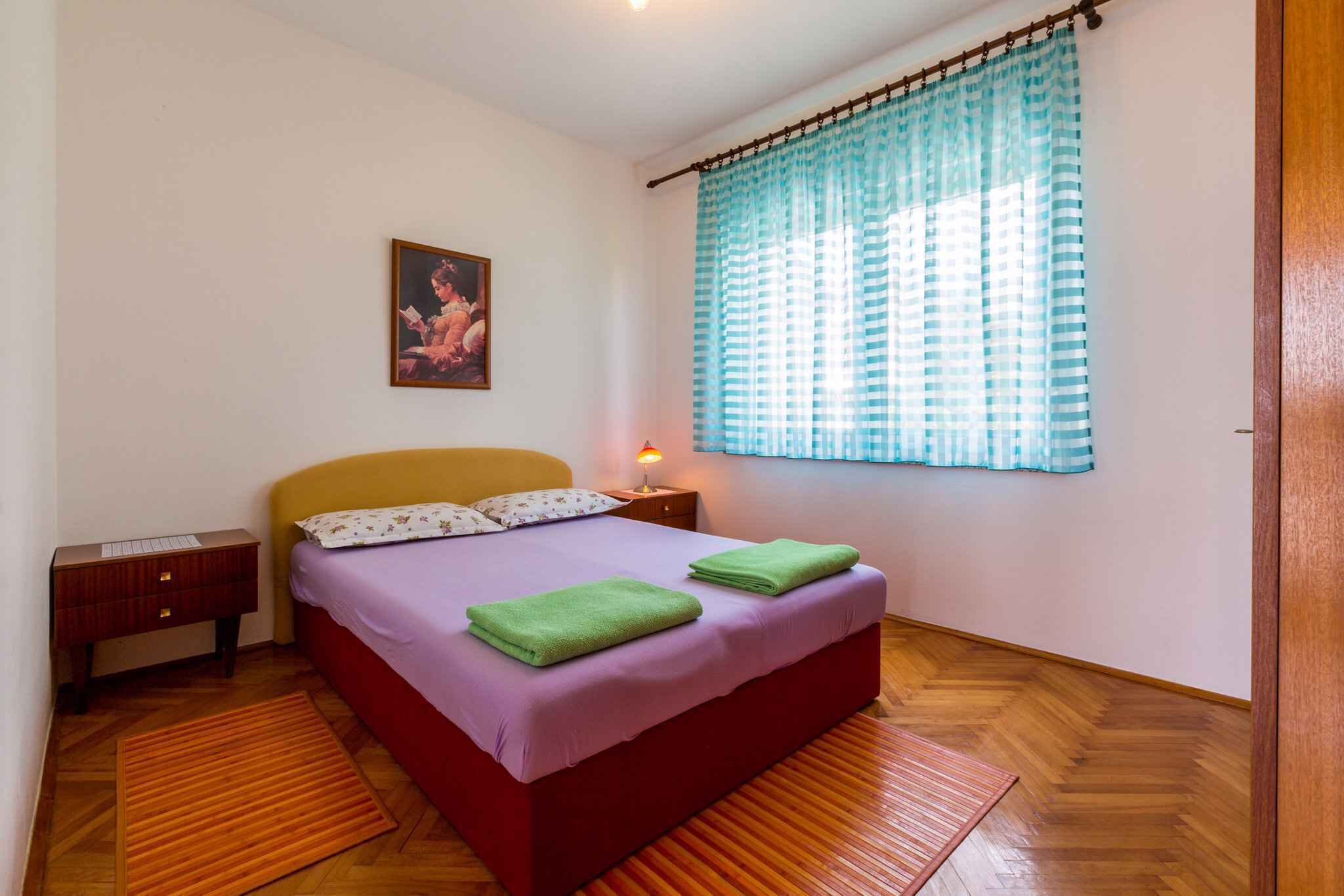 A bedroom with a bed, a nightstand, a lamp, a painting, and a window with curtains.