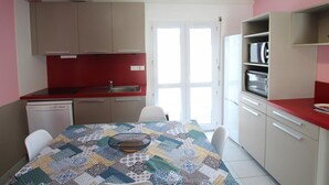 Cocina compacta con encimera roja, lavamanos y lavavajillas. Se aprecia una mesa con mantel estampado y sillas.