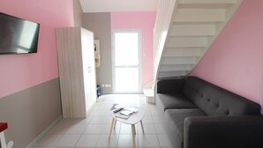 Una sala de estar moderna con un sofá gris, una pequeña mesa blanca con libros y un control remoto, un televisor montado en la pared y una escalera que lleva a un nivel superior.