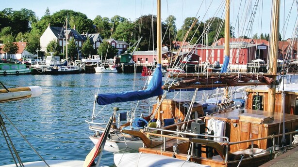 Un port de plaisance avec plusieurs bateaux amarrés, des bâtiments en bois en arrière-plan et un ciel dégagé.