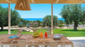 An outdoor dining area with a wooden table, white chairs, and a view of the sea. - Casa Carol - Blu - Independent Villa Modica Area (Scicli)
