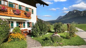 Ein zweistöckiges Gebäude mit einem Blumen geschmückten Balkon, einem steinernen Weg, der zum Eingang führt, und einer gebirgigen Kulisse.