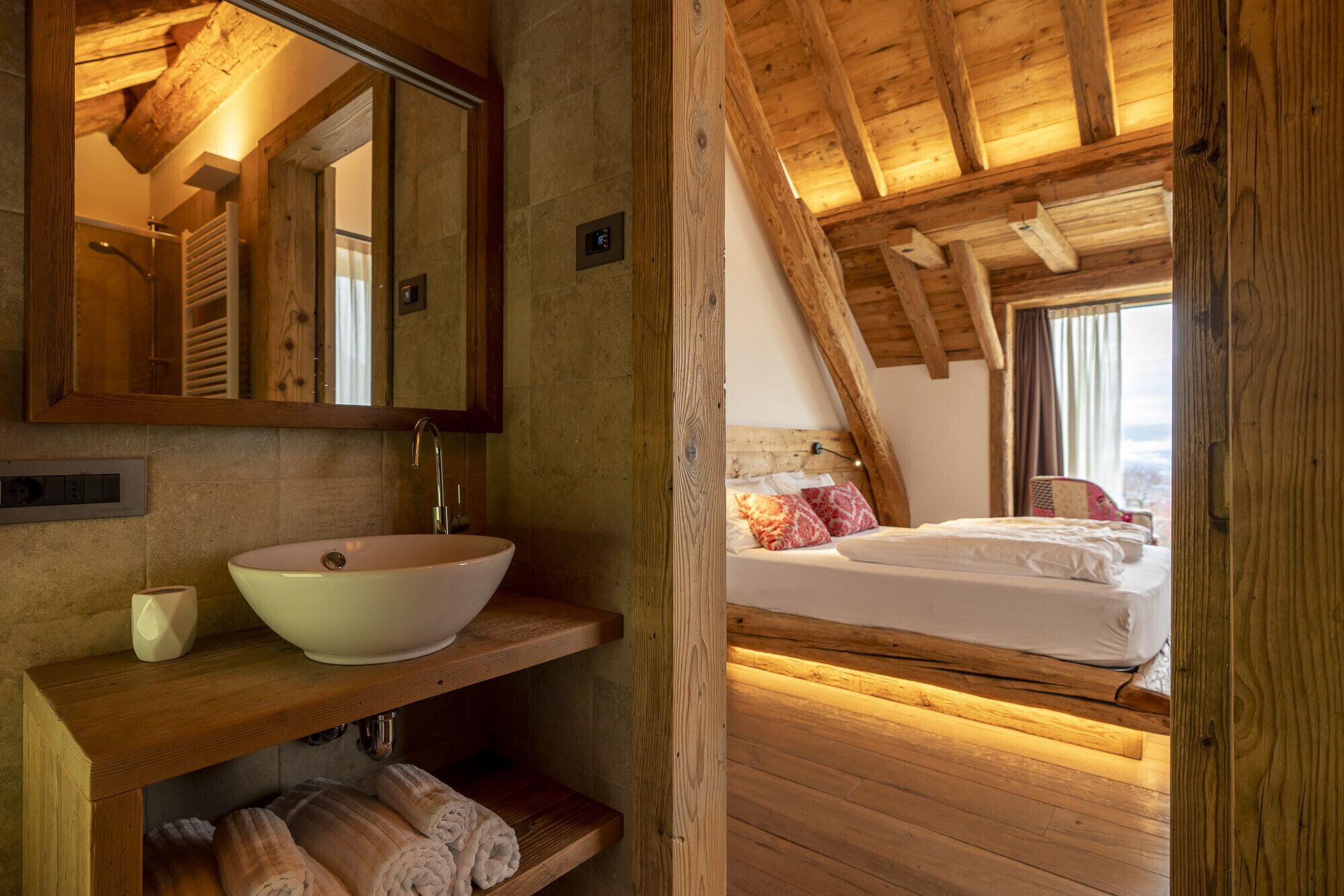 Une chambre à coucher rustique avec un plafond en bois, une grande fenêtre, un lit avec des oreillers et une table de chevet en bois surmontée d’un lavabo en forme de coupe.