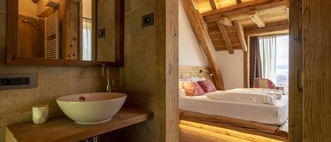 Une chambre à coucher rustique avec un plafond en bois, une grande fenêtre, un lit avec des oreillers et une table de chevet en bois surmontée d’un lavabo en forme de coupe.