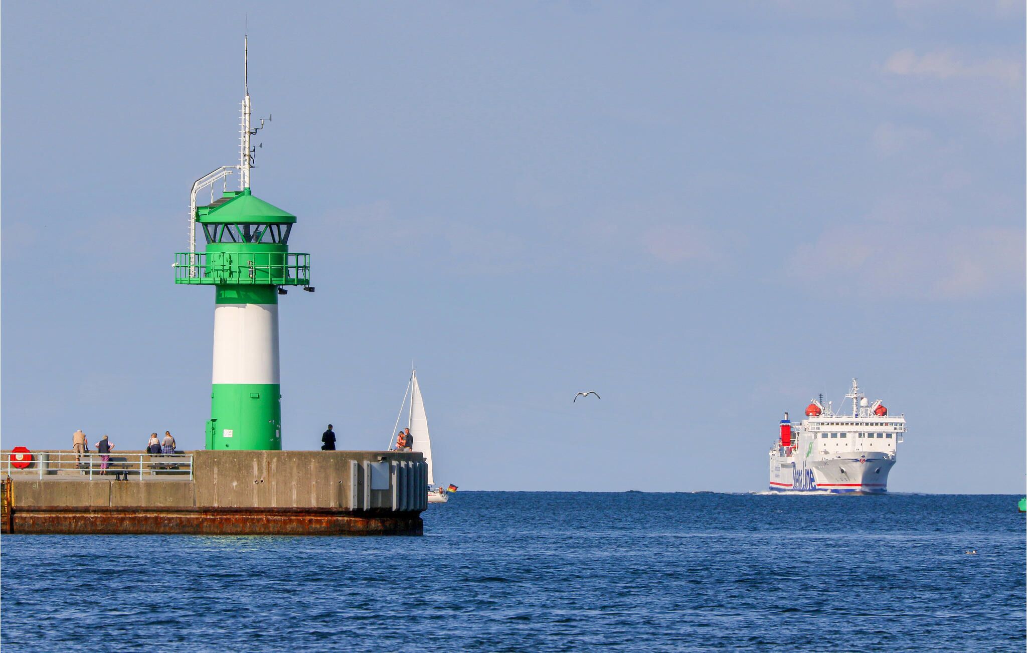 A lighthouse with a green and white structure, a pier with people, and a large ship in the background.