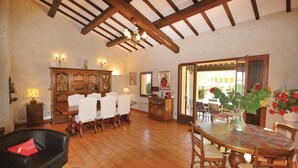 Uma ampla área de jantar com teto alto e vigas aparentes, uma mesa de jantar com cadeiras, um sofá de couro preto e um armário de madeira com portas de vidro.