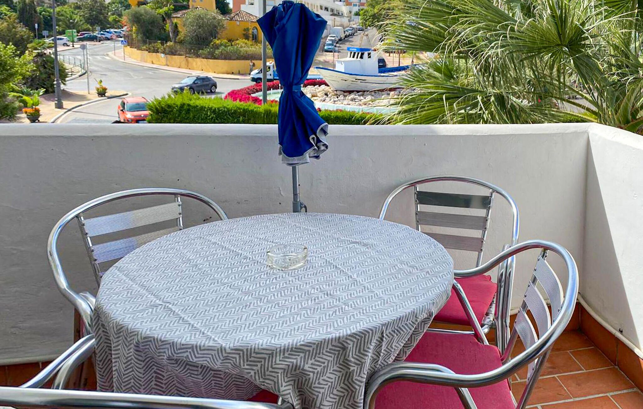 Un balcón con una mesa redonda y sillas, una sombrilla azul y vistas a una calle con coches y edificios.