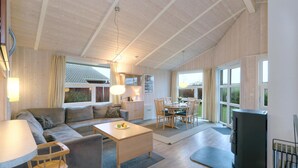 A spacious living area with a sofa, coffee table, and dining table. There is a fireplace and a large window providing natural light. - Friedrichskoog-Strandpark 18 (Friedrichskoog)