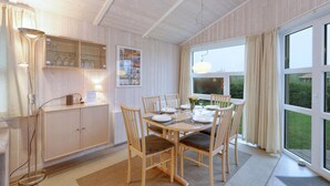A dining area with a wooden table set for four, surrounded by chairs. There is a cabinet with glassware and a lamp to the left. A large window provides a view of the outdoors. - Friedrichskoog-Strandpark 18 (Friedrichskoog)