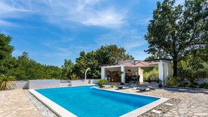 Ein rechteckiger Pool mit Steinbordstein, eine überdachte Außenlounge mit Liegestühlen und eine Pergola.