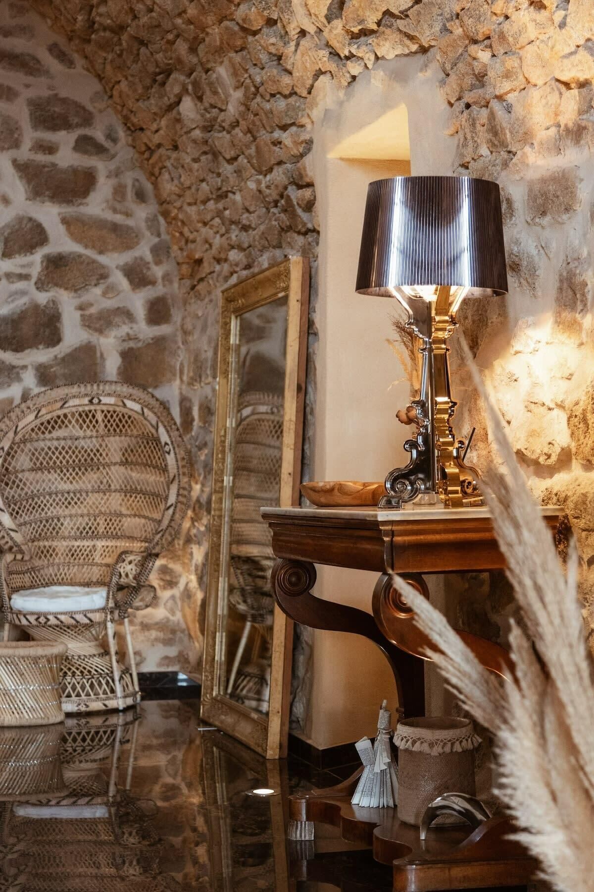 A room with a stone wall, a wooden table with a lamp, a mirror, and wicker furniture.