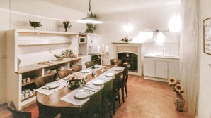 Un comedor con una larga mesa dispuesta para una comida, rodeada de sillas. Hay estanterías con utensilios de cocina y una planta pequeña en la pared.