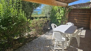 Un patio exterior con una pérgola de madera, suelo de piedra y sillas de plástico blanco.
