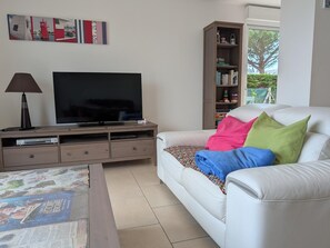 Ein Wohnzimmer mit einem Flachbildfernseher auf einem Holzboden, ein weißes Sofa mit bunten Kissen, einem Bücherregal und einem Fenster mit Blick auf Bäume.