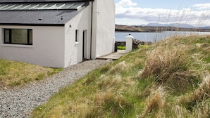 A white house with a sloped roof and skylights, a gravel path leading to the entrance, and a view of a lake with mountains in the distance. - 3 bedroom accommodation in Roag, Isle of Skye (Roag, Isle of Skye)