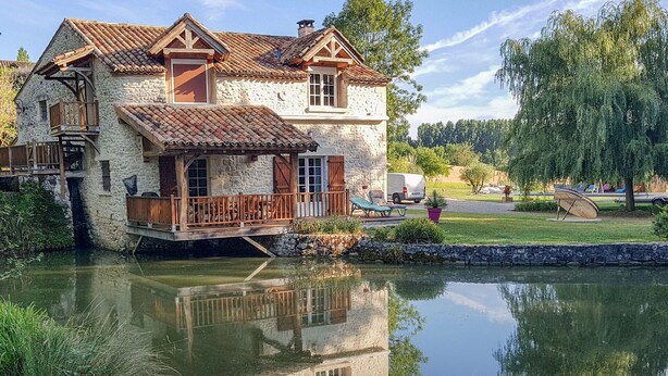 Una casa de piedra con balcón de madera, un estanque en primer plano y el reflejo de la casa en el agua.