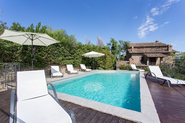A rectangular swimming pool with white lounge chairs and umbrellas, surrounded by a wooden deck and stone walls.