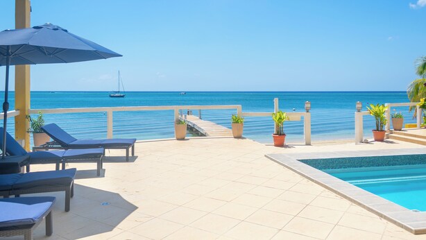 Área da piscina com espreguiçadeiras e um guarda-sol, com vista para um mar azul e límpido onde se avista um veleiro ao longe.