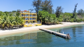 Um hotel amarelo de vários andares com varandas, uma praia e um cais que se estende para a água.