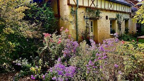 Una casa tradicional con techo a dos aguas, una chimenea visible y un jardín con diversas plantas florecientes.