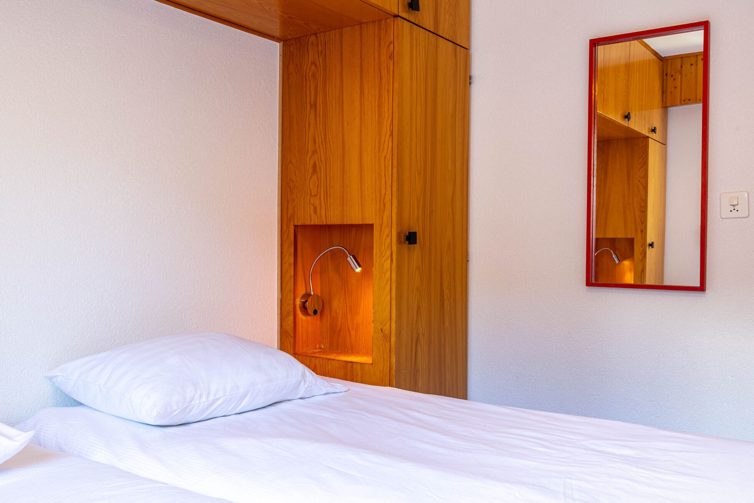 A bedroom with a wooden wardrobe, a bed with white bedding, and a mirror with a red frame.
