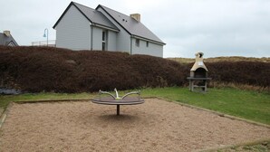 A house with a grey roof and white walls, a gravel area with a metal sculpture, and a hedge in the background.