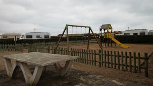 A playground with a swing set and slide, surrounded by a wooden fence, with caravans in the background.