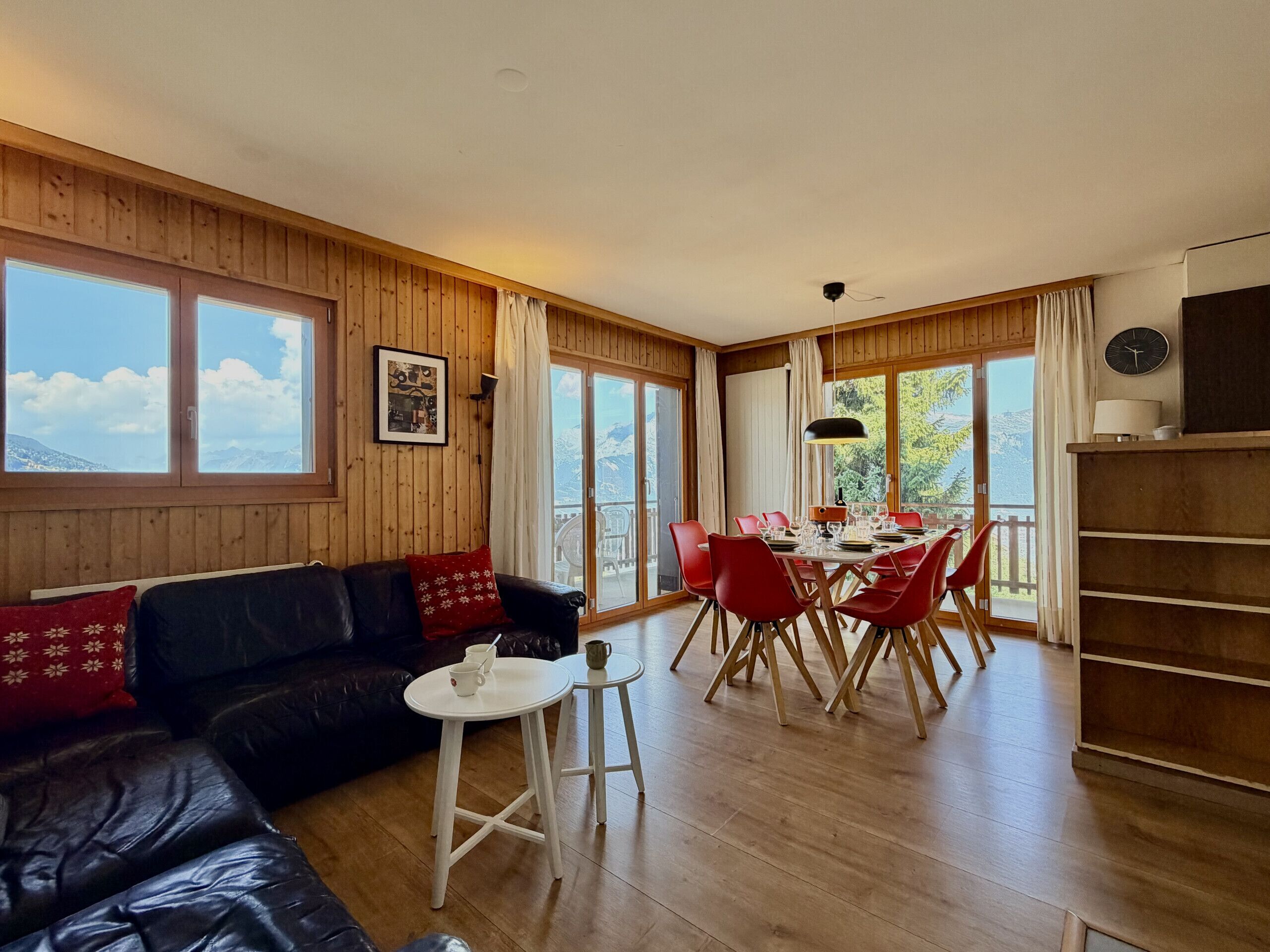 Ein Zimmer mit Holzvertäfelung, einem schwarzen Ledersofa, einem Esstisch mit roten Stühlen und großen Fenstern.