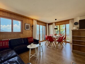 A room with a wooden interior, a black leather sofa, a dining table with red chairs, and large windows. - Gentianes G 010 - COSY apartment 8 pers (Veysonnaz)
