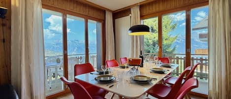 Ein Essbereich mit einem Tisch für sechs Personen, roten Stühlen, einer schwarzen Pendelleuchte und großen Fenstern mit Blick auf die Berge.