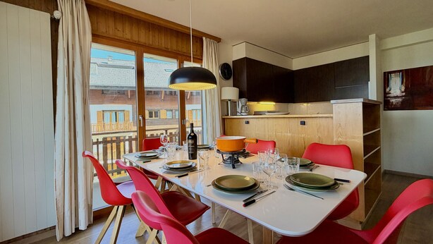 A dining area with a table set for four, red chairs, a black pendant light, and a view of a balcony with wooden railings. - Gentianes G 010 - COSY apartment 8 pers (Veysonnaz)