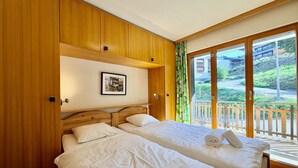 A hotel room with two beds, wooden paneling, a framed picture on the wall, and a view of a balcony with a wooden railing. - Gentianes G 010 - COSY apartment 8 pers (Veysonnaz)