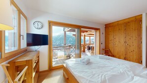 A bedroom with a bed, a TV, a wooden desk, a wardrobe, and a sliding glass door leading to a balcony with a mountain view.