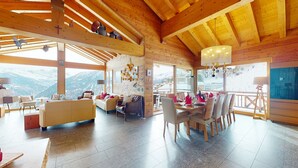 A spacious room with a high ceiling and wooden beams, furnished with leather sofas and a dining table set for six. Large windows offer a view of snow-capped mountains.